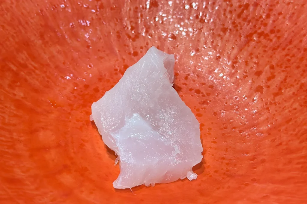 A single, clean slice of translucent white fish sashimi served in a textured orange ceramic bowl, highlighting its fresh texture.