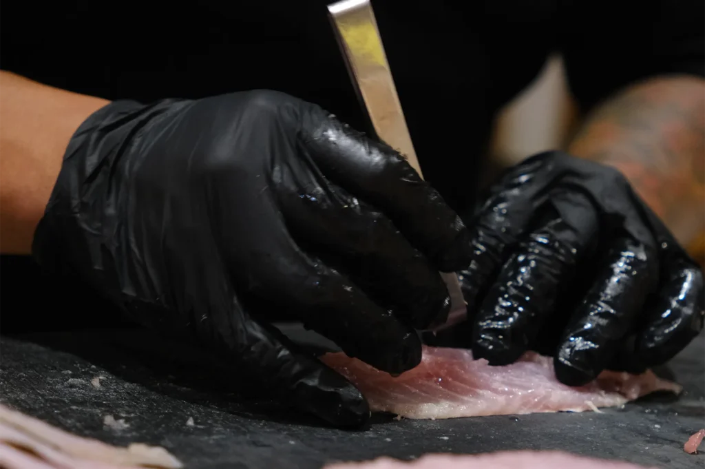 Close-up of a chef wearing black gloves using professional fish bone tweezers to prep a white fish fillet on a dark stone board.