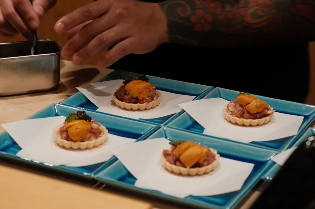 Four delicate savory tartlets topped with minced tuna tartare, fresh uni, and caviar, being prepared by a chef in a fine-dining setting.