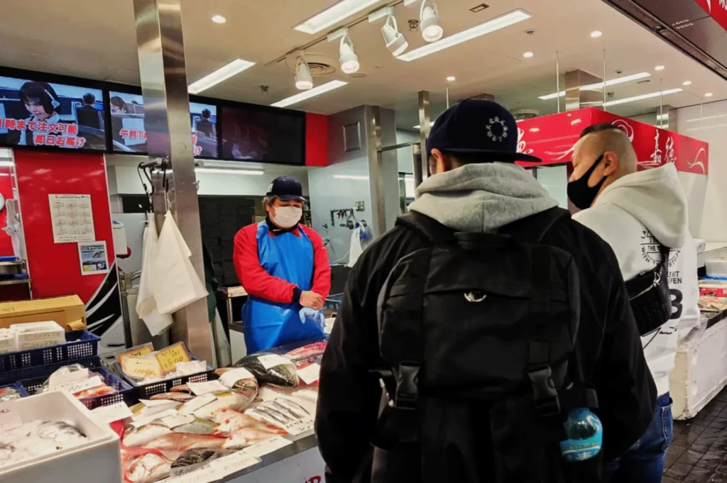 Two men wearing hoodies and masks stand at a fish market counter. The vendor, in a red and blue uniform and mask, prepares fish for them.