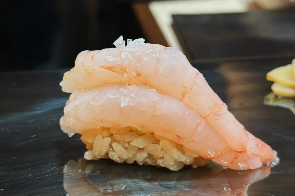 Close-up of a sushi piece with translucent, pink shrimp atop rice, garnished with clear salt crystals, on a metal surface. Elegant and fresh presentation.