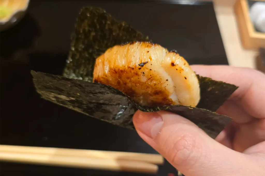 A hand holding a large, perfectly seared hotate (scallop) wrapped in a crisp sheet of toasted nori seaweed.