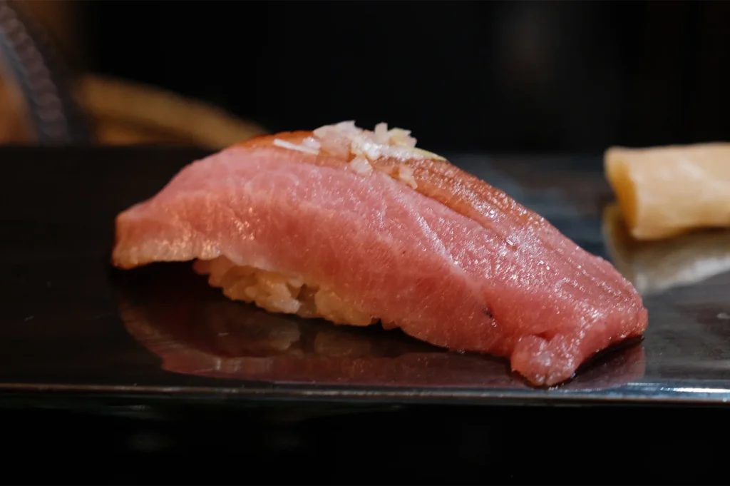 A piece of Otoro (fatty tuna) nigiri sushi with a light soy glaze and garnish, resting on a black lacquered sushi board.