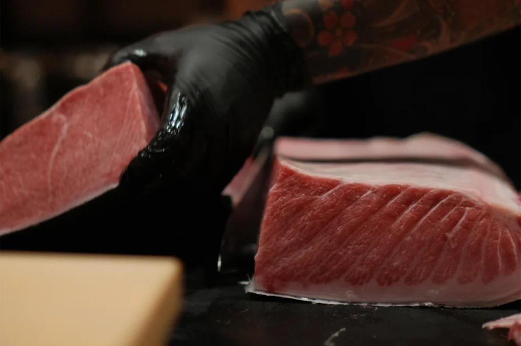 A chef with a traditional tattoo slicing a large, highly marbled block of bluefin tuna belly (Otoro) with a sharp sushi knife.