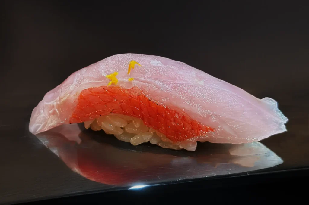 A piece of sushi with glossy rice topped by a slice of pink and red fish, garnished with tiny yellow zest, placed on a reflective surface.