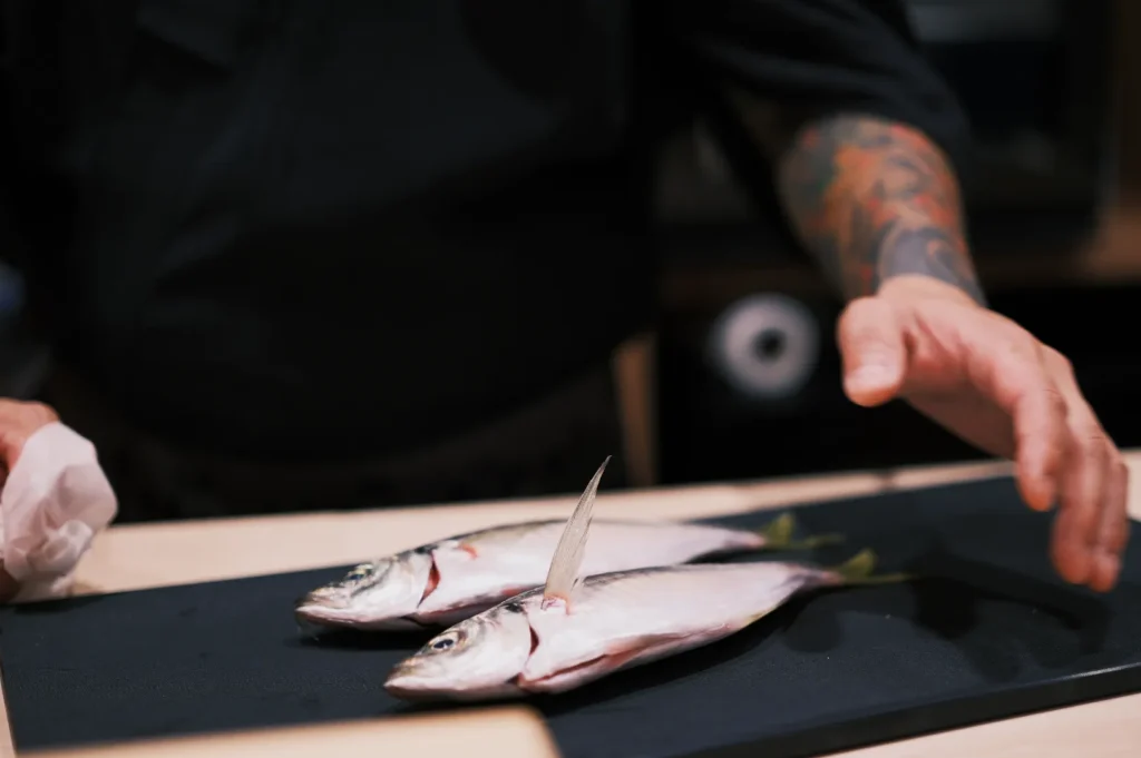 Chef with a tattooed arm prepares two fresh fish on a black cutting board. The scene suggests focus and culinary expertise.