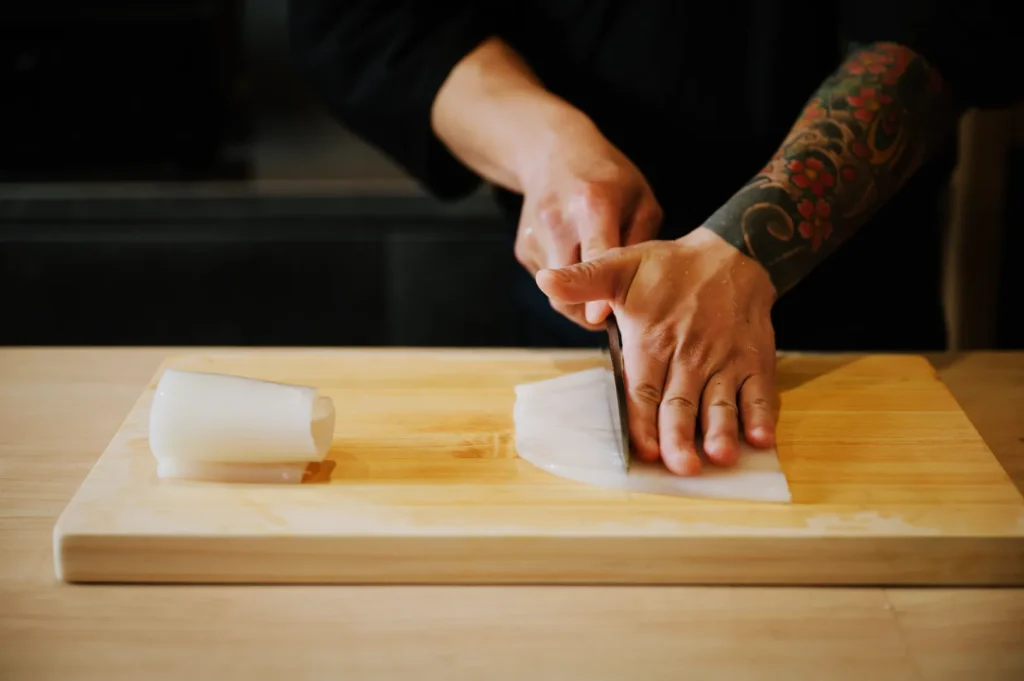 A person with tattooed arms skillfully slices a piece of squid on a wooden cutting board in a kitchen, conveying precision and focus.