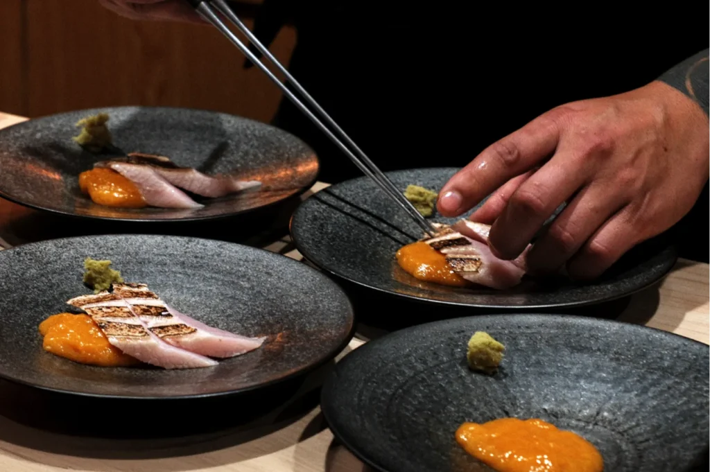 A chef meticulously arranges grilled fish slices with orange sauce and green garnish on black plates, showcasing culinary precision and elegance.
