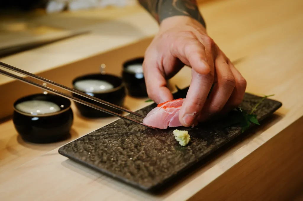 A hand artistically places a slice of sushi on a textured black slate plate with chopsticks. Wasabi and green leaves accent the minimalist sushi presentation, creating a refined and elegant atmosphere.