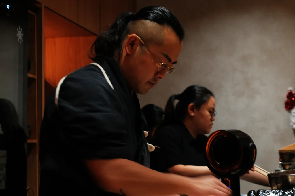 Two chefs focus intently on their tasks in a warmly lit kitchen. One is wearing glasses, and both are clad in dark clothing, creating a calm, professional atmosphere.