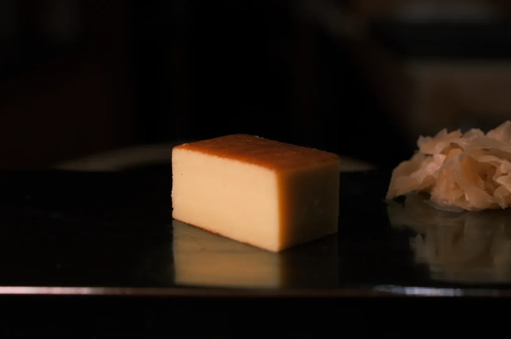 A slice of dense, golden-brown cake sits on a reflective black surface, accompanied by a small mound of pickled ginger, creating a minimalist and elegant presentation.