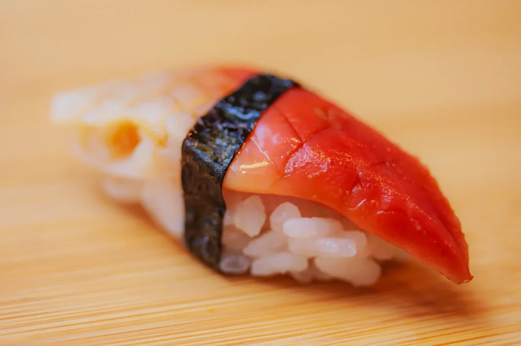 Close-up of a sushi piece with red tuna and creamy octopus, wrapped in nori seaweed on rice, placed on a light wooden surface. Fresh and inviting.