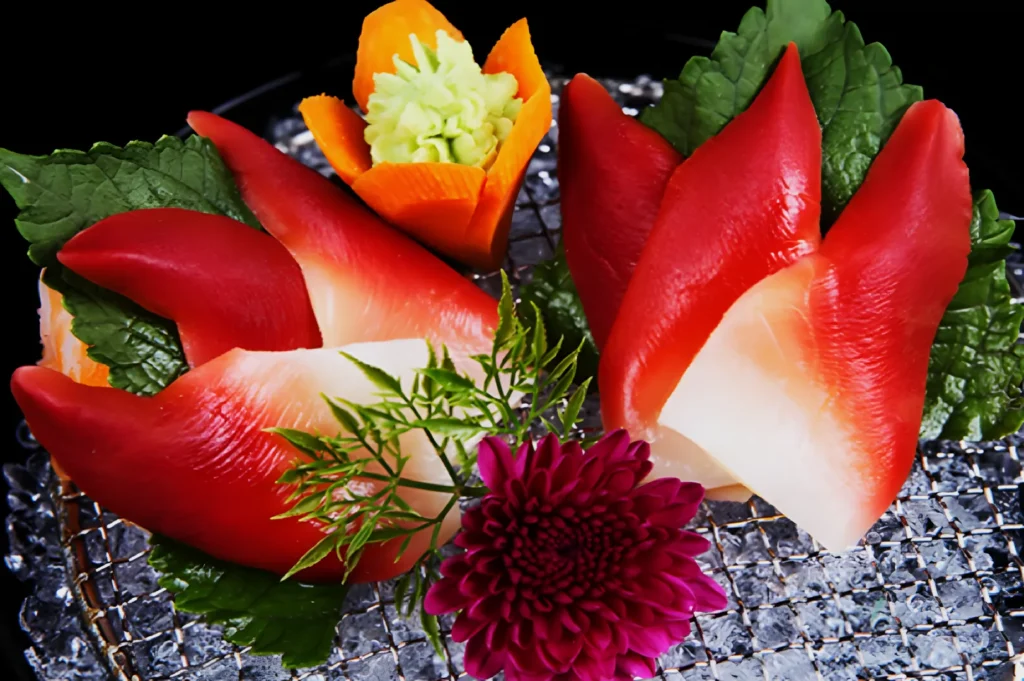 Close-up of a vibrant sushi platter featuring red clam sashimi on green shiso leaves, garnished with a purple chrysanthemum and carved carrot flower.