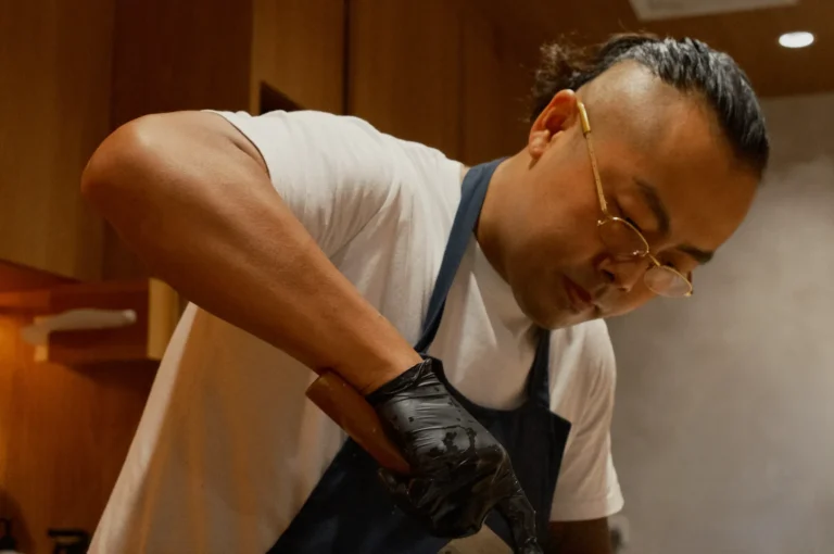 Chef Masa focused on slicing food, wearing glasses, a white shirt, and apron. He has short hair tied in a bun and wears black gloves in a kitchen setting.