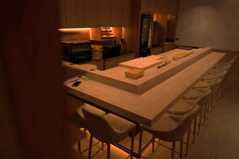 Warmly lit sushi bar with wooden counters and a dozen cushioned chairs. Kitchen tools on the counter suggest a cozy, inviting atmosphere.