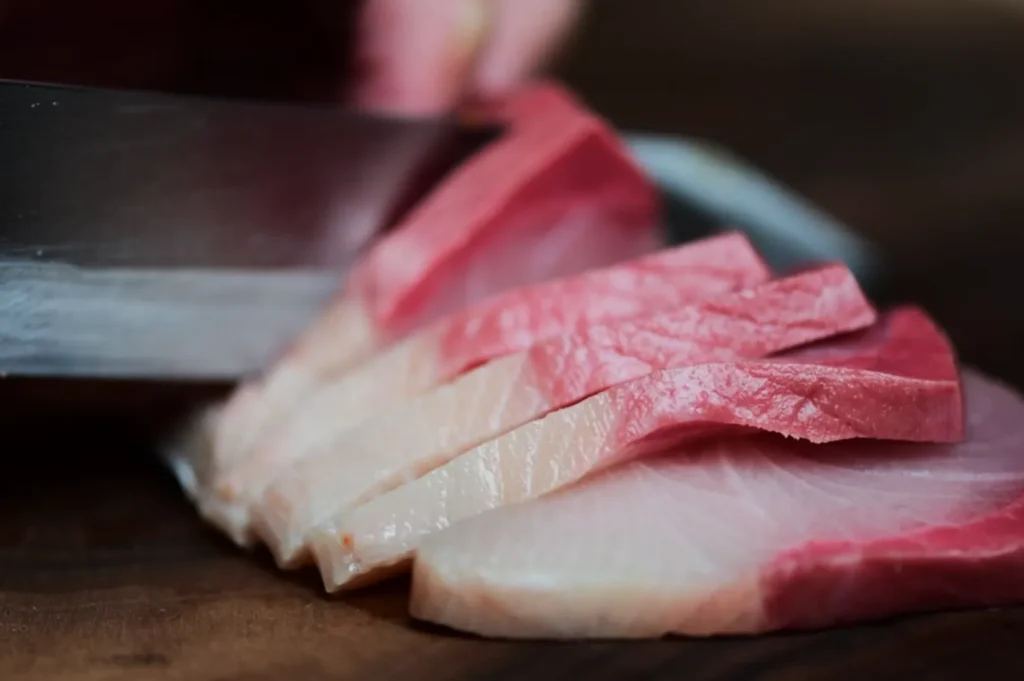 Close-up of a knife slicing raw fish with a marbled texture and pinkish hue on a wooden board, conveying freshness and precision in preparation.