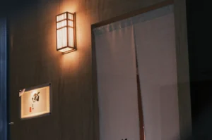A warm-lit, Japanese-style entrance with a wooden wall, featuring a vertical rectangular lantern and noren curtain. A sign with kanji is displayed nearby.