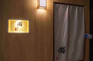 Elegant wooden entrance with Japanese curtain and illuminated sign featuring calligraphy. Soft lighting creates a calm, inviting atmosphere.