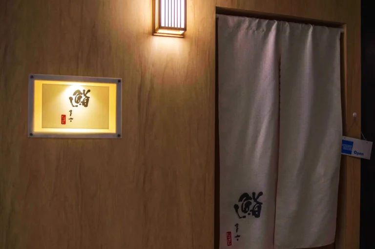 Elegant wooden entrance with Japanese curtain and illuminated sign featuring calligraphy. Soft lighting creates a calm, inviting atmosphere.