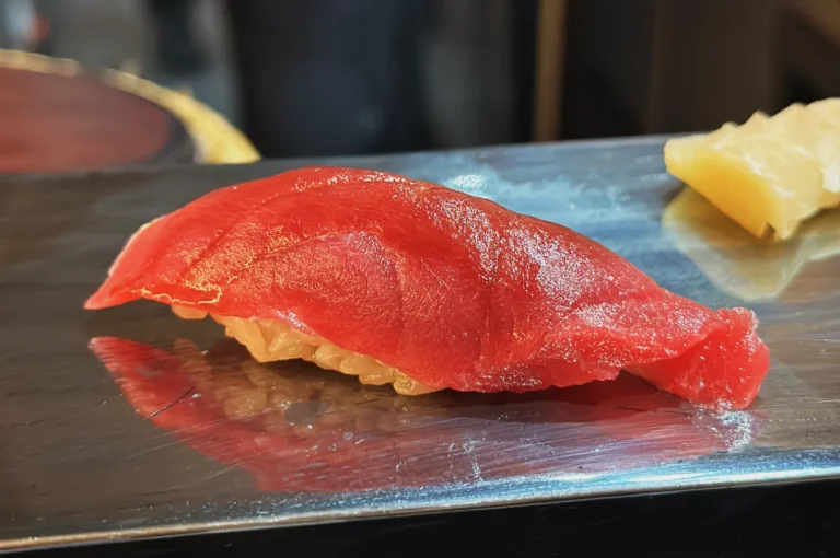 Close-up of a piece of nigiri sushi with a glossy red tuna slice on top of vinegared rice, placed on a reflective silver plate, creating a fresh and appetizing look.