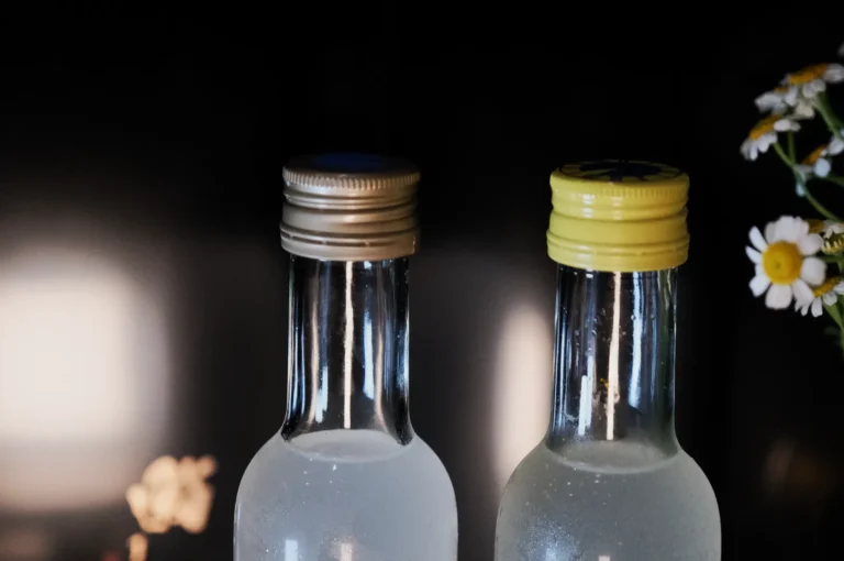 Two clear glass bottles with metal caps, one gold and one yellow, sit on a dark background. White flowers bloom on the right, adding a fresh touch.