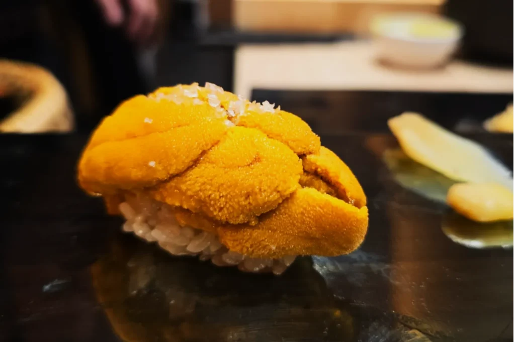Close-up of a sushi piece topped with vibrant orange uni (sea urchin) on rice, set on a glossy black plate with a slice of pickled ginger beside it.
