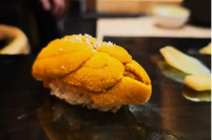 Close-up of a sushi piece topped with vibrant orange uni (sea urchin) on rice, set on a glossy black plate with a slice of pickled ginger beside it.