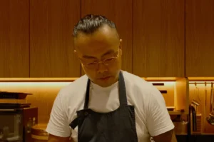 A person in a white t-shirt and black apron stands focused in a warmly lit kitchen with wooden cabinets, conveying a calm, professional ambiance.