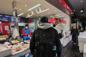 Two people shopping at a fish market stall, with a vendor in a mask and apron behind the counter. Displayed fish are visible. Indoor setting with bright lighting.
