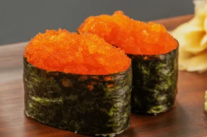 Close-up of two sushi pieces topped with bright orange roe, wrapped in dark green seaweed. Set on a wooden surface, conveying a fresh, savory vibe.