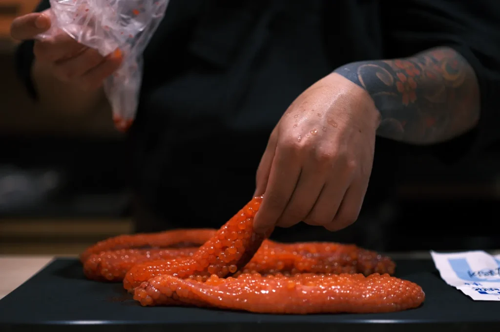 Close-up of a person with tattooed arm preparing bright orange salmon roe on a black surface. The setting is a dimly lit kitchen, creating a focused, detailed atmosphere.