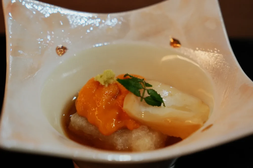 A delicate Japanese dish in a white textured bowl features a vibrant orange sea urchin, a slice of white seafood, a green herb garnish, and sauce, exuding elegance.