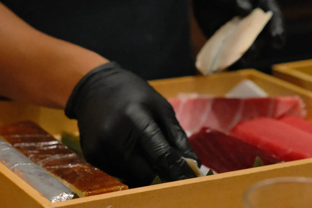 A person wearing black gloves is selecting sushi from a wooden box, featuring colorful fish slices. The scene conveys a meticulous, culinary focus.