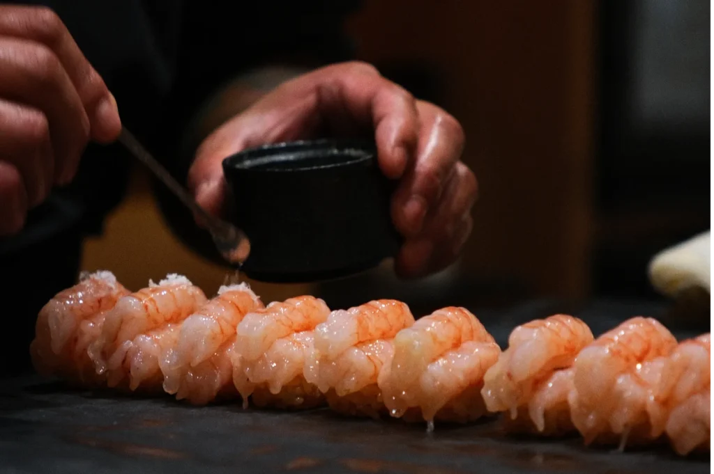 A chef's hands are delicately seasoning shrimp sushi with a spoonful of sauce. The sushi is neatly aligned, conveying precision and craftsmanship.