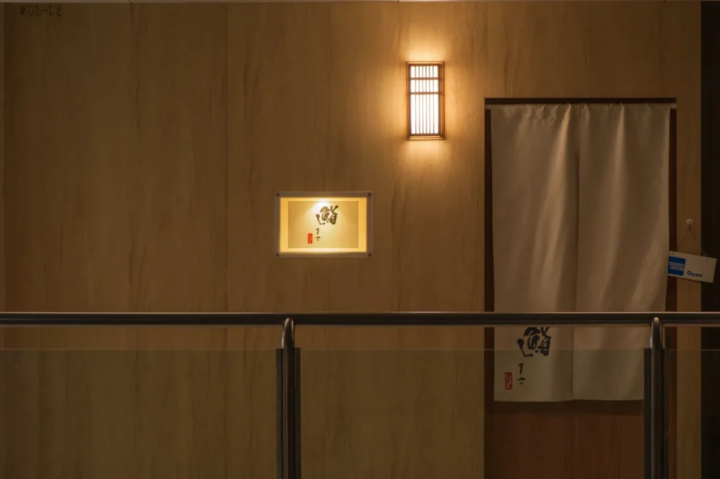 Dimly lit wooden hallway with a glowing wall sconce, a simple curtain covering a doorway, and a small illuminated sign with Japanese characters. Calm ambiance.
