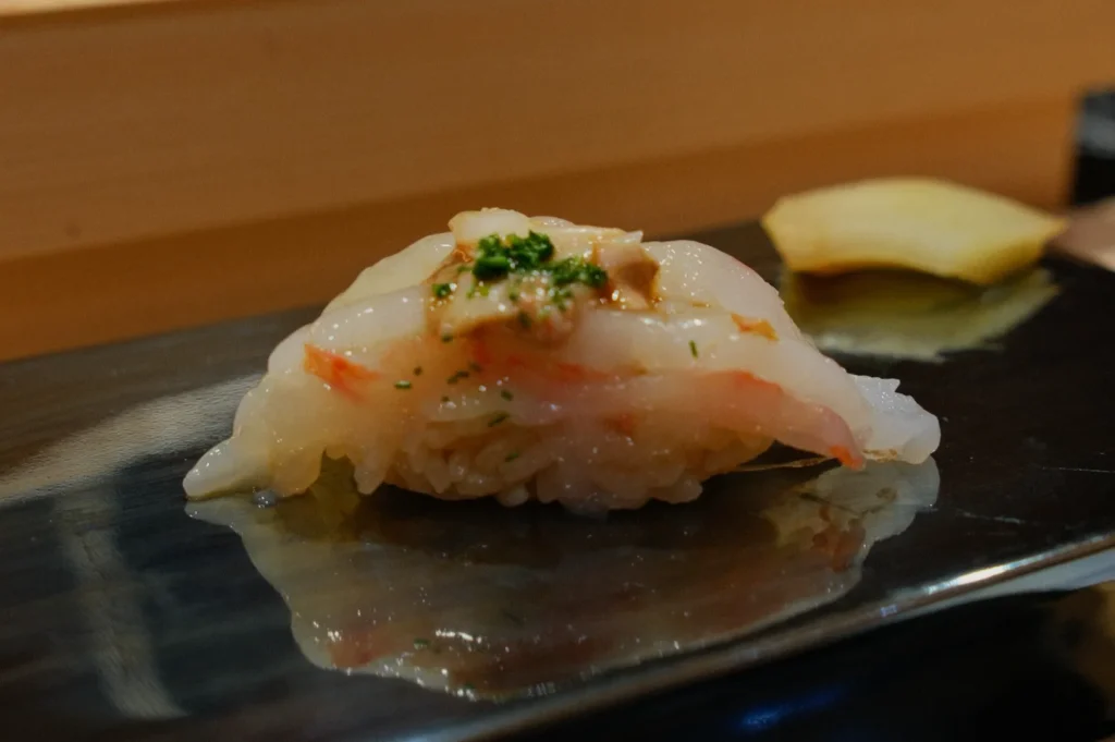 Close-up of a piece of sushi on a glossy black plate. The sushi is topped with delicate white fish, a sprinkle of green herbs, and garnished with a small, light brown topping, creating a refined and appetizing appearance. A slice of yellow pickled ginger is blurred in the background, suggesting a serene dining experience.