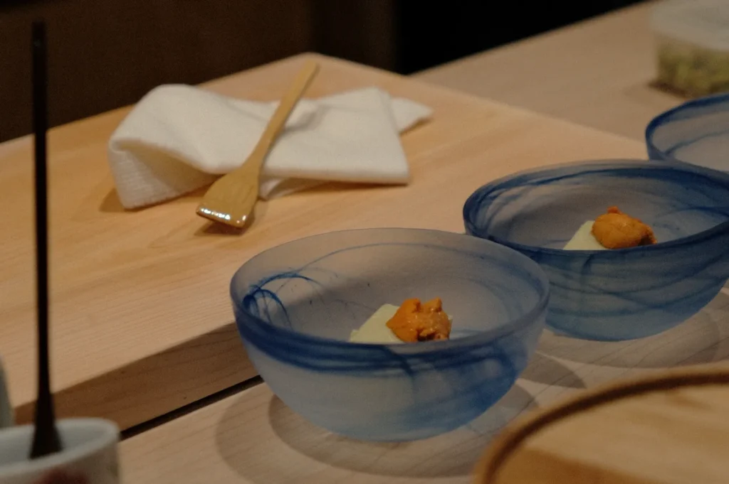 Two blue glass bowls, each with sea urchin on a cream base, rest on a wooden table. A folded white napkin and wooden spoon are placed nearby, creating an elegant dining setting.