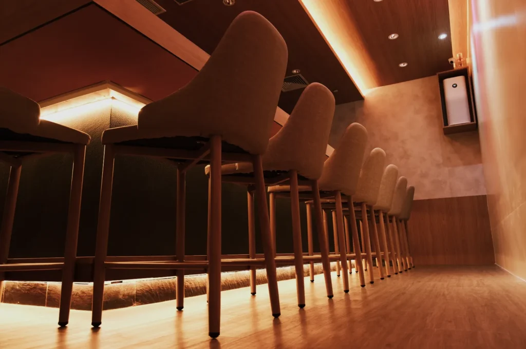 Bar interior with a row of upholstered chairs and wooden legs, dimly lit by warm ceiling lights. The atmosphere is cozy and inviting.