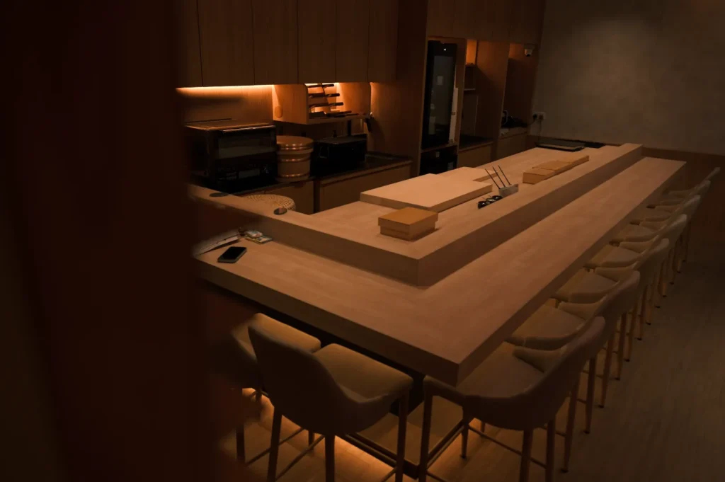 Dimly lit, minimalist sushi bar interior with wooden counters and chairs. Soft lighting creates a warm, inviting atmosphere. Empty seats line the counter.