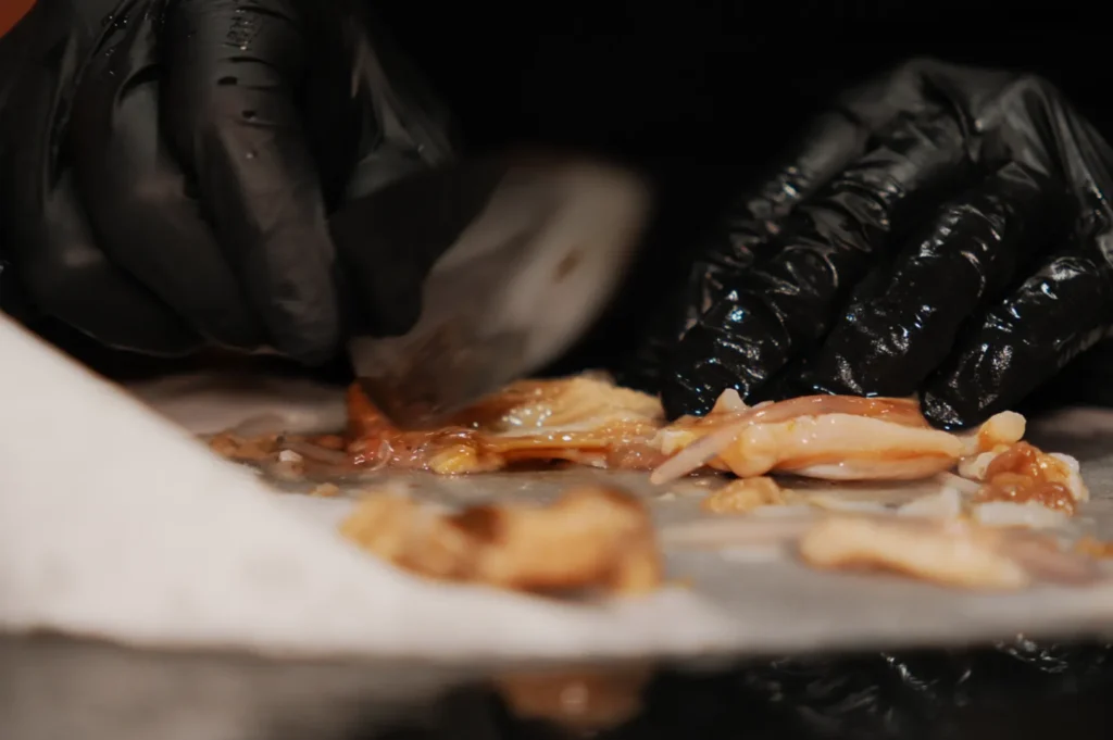 Hands in black gloves skillfully slice raw chicken on a cutting board with a silver knife, conveying focus and precision in food preparation.