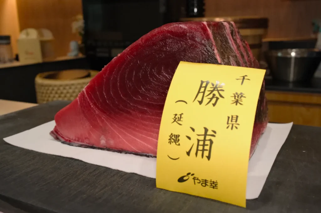 A thick, fresh cut of deep red tuna with a visible texture sits on a white surface. A yellow label with Japanese text is attached to the fish. The background features a kitchen setting with dim lighting, evoking a sense of culinary craftsmanship.