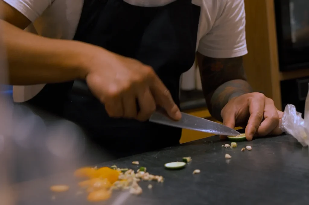 A person wearing an apron slices a cucumber on a kitchen counter, surrounded by chopped vegetables. The scene conveys focus and culinary skill.