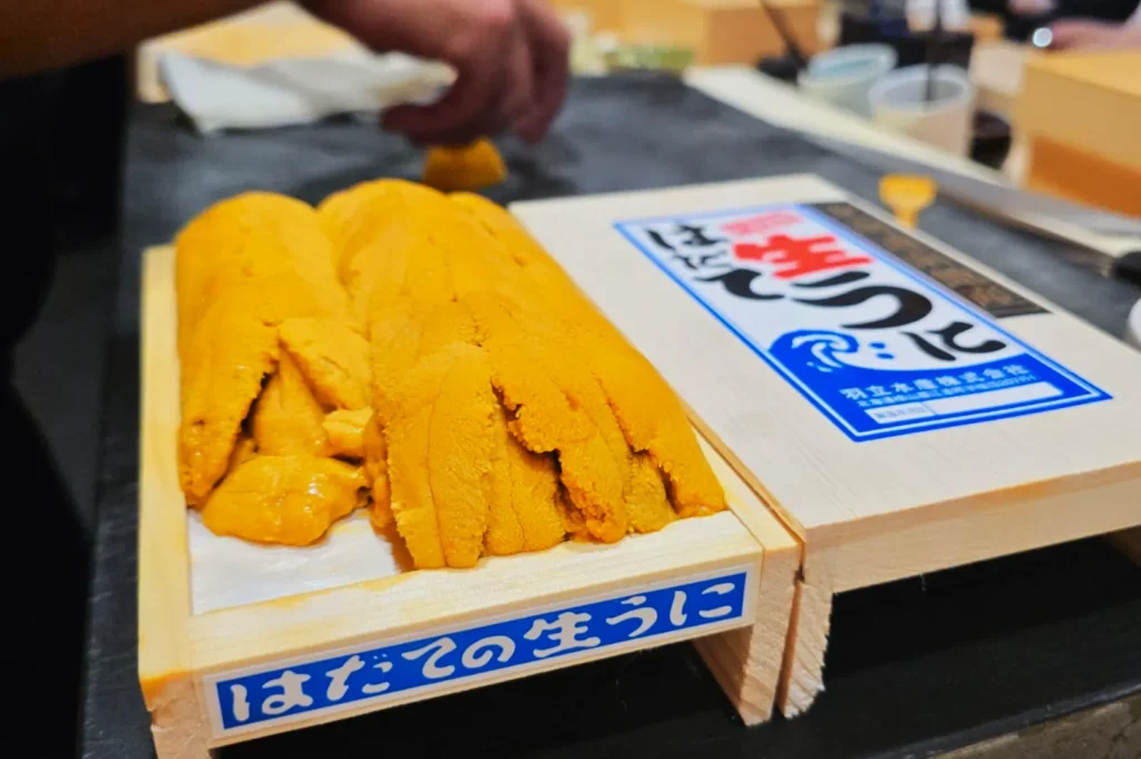 A wooden tray filled with fresh, bright orange sea urchin (uni) is placed on a counter. A hand reaches out in the background, suggesting preparation. The setting conveys a culinary, artisanal atmosphere.