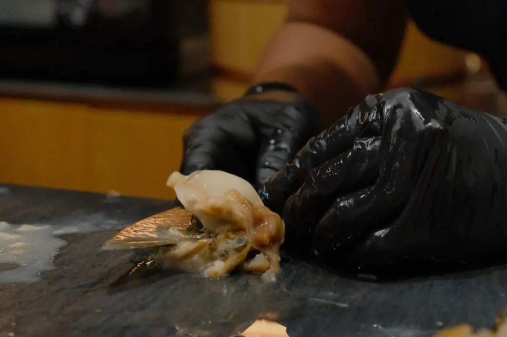 Hands in black gloves shuck an oyster on a dark countertop. The scene conveys a sense of skill and focus in a kitchen setting.