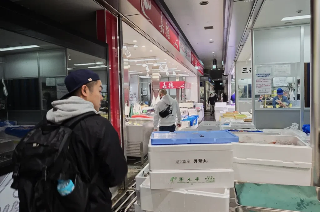 A man wearing a backpack and cap walks through a bustling fish market with overhead lights. Vendors and seafood displays create a lively, busy atmosphere.