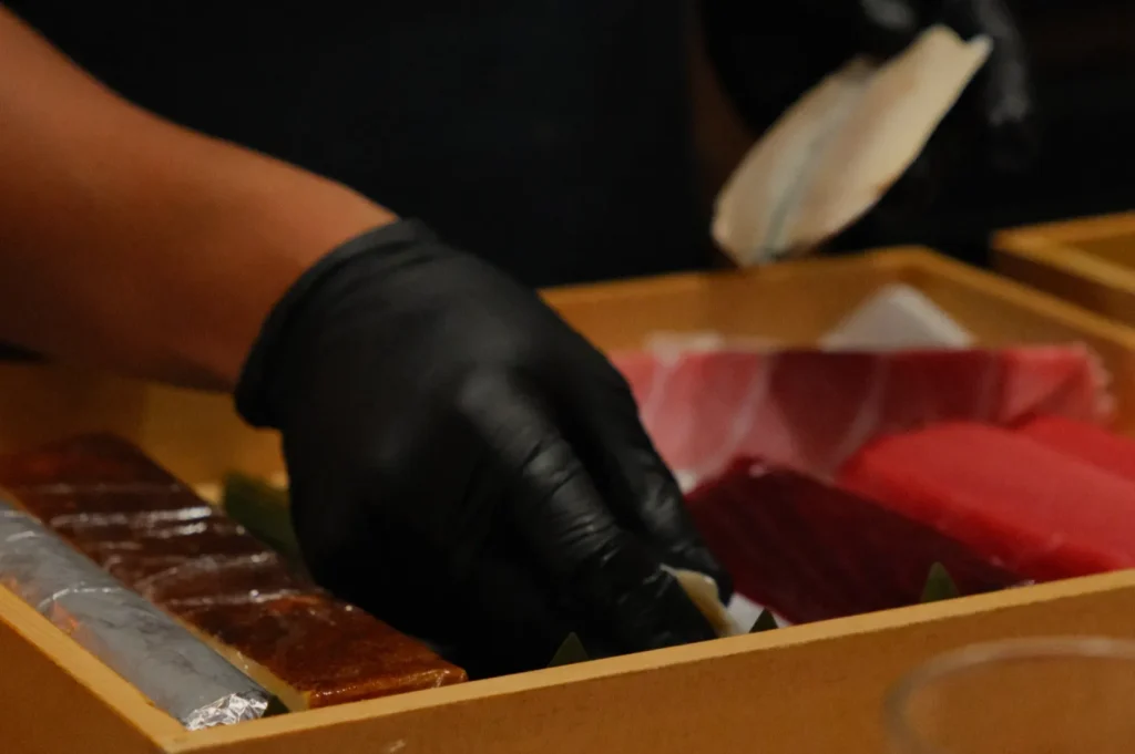 A person wearing black gloves prepares sushi, placing slices of raw fish into a wooden box. The scene conveys skill and focus in sushi making.