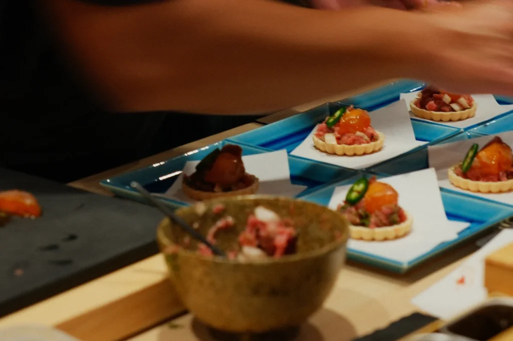 A chef's hands arrange appetizers on blue trays, showcasing vibrant tartlets with diced meat, a glossy topping, and green garnish, conveying precision and artistry.
