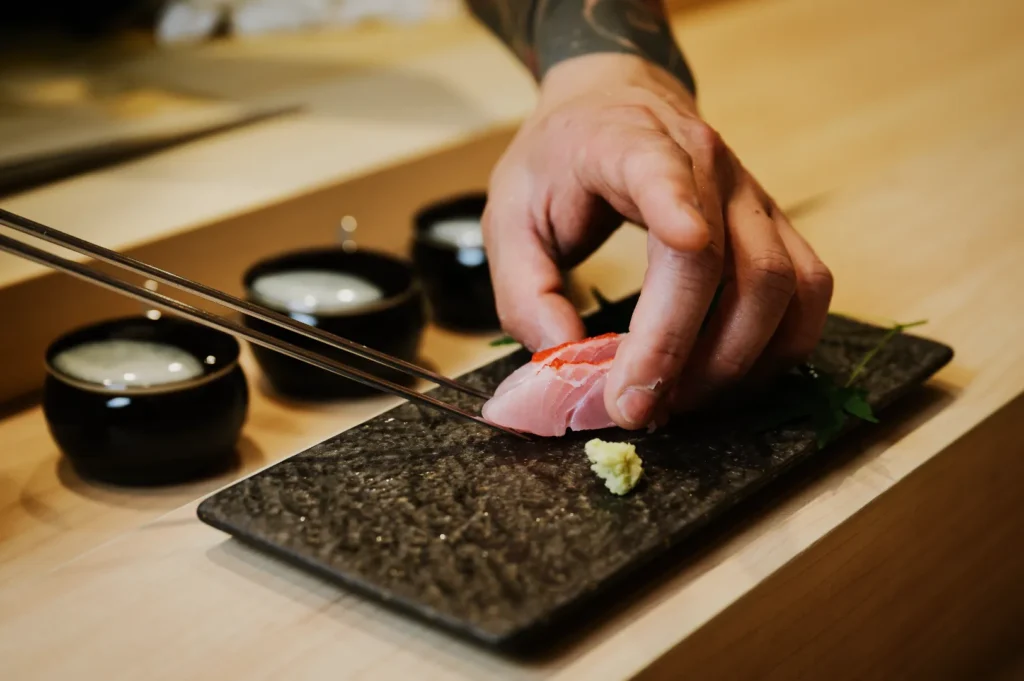 A hand with chopsticks places a slice of sashimi on a textured stone plate beside wasabi and greens, with small black bowls in the background.