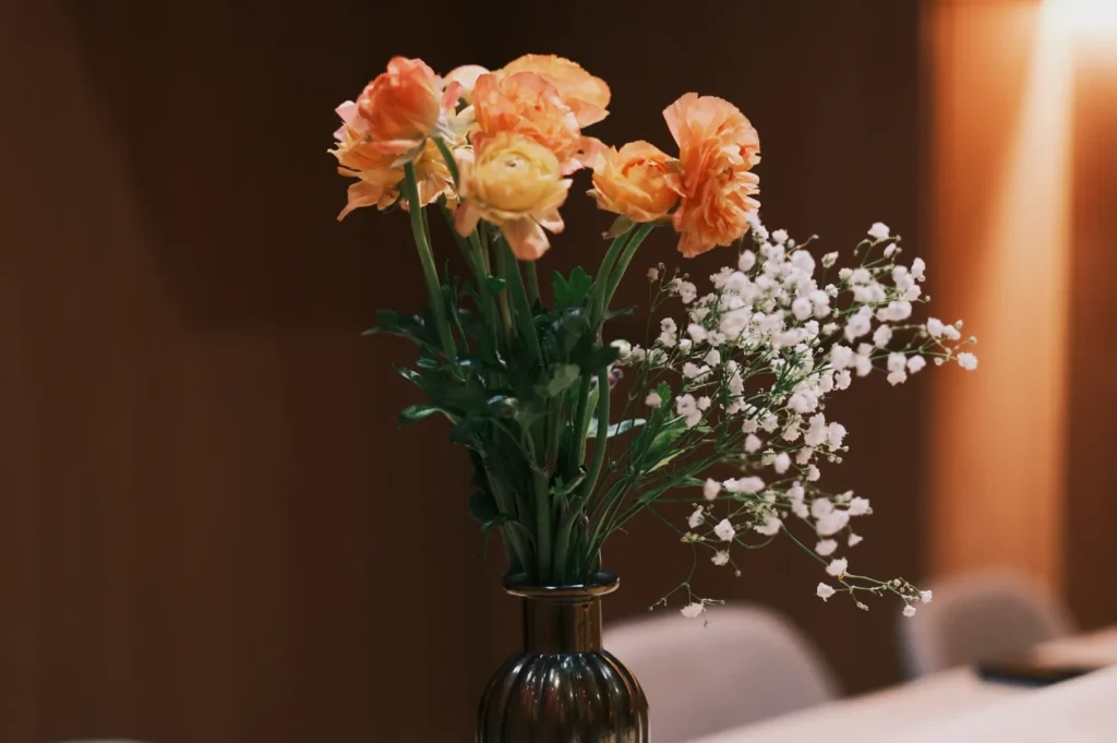 A bouquet of orange flowers and white baby's breath in a dark vase on a wooden table. The soft lighting and blurred background create a serene, cozy ambiance.