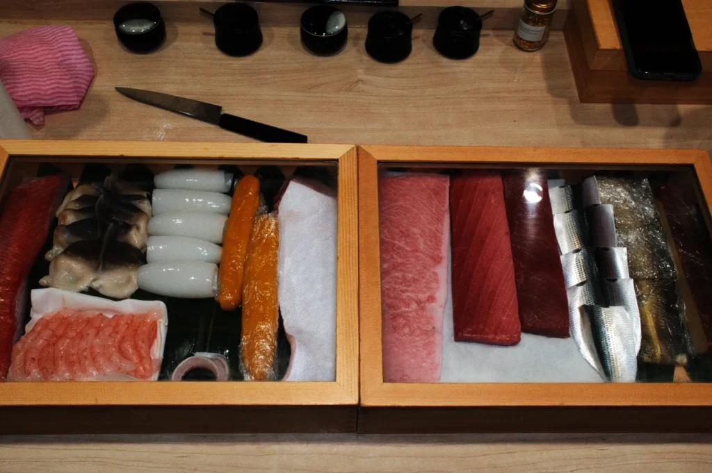 A variety of fresh sushi ingredients in two wooden display boxes on a counter. Items include fish and roe. A knife rests nearby on the wooden surface.